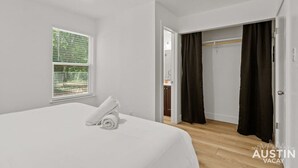 Une chambre à coucher avec un lit, une fenêtre avec des stores, un dressing avec un miroir et une porte donnant sur une autre pièce.