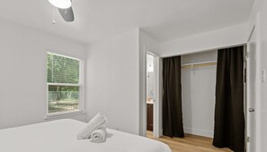 Une chambre à coucher avec un lit, une fenêtre avec des stores, un dressing avec un miroir et une porte donnant sur une autre pièce.