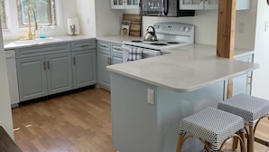 A modern kitchen with light gray cabinets, a central island with stools, a microwave, and a kettle. - Newly Renovated Quonnie Beach House -  QEBA Beach Pass & Pond Access (Charlestown)