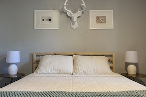 A bedroom with a bed, two bedside tables, a wall-mounted deer head, and two framed pictures.