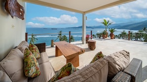 A spacious balcony with a view of the ocean, furnished with a sofa, a wooden table, and potted plants. - Oceanview 4BR Villa,Infinity Pool in East Bali (Candidasa)