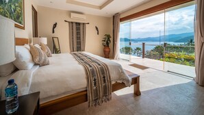 A bedroom with a large bed, a bedside table with a water bottle, a potted plant, and a view of the sea through a glass door. - Oceanview 4BR Villa,Infinity Pool in East Bali (Candidasa)
