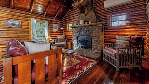 A log cabin interior with a stone fireplace, wooden furniture, and a mounted deer head.