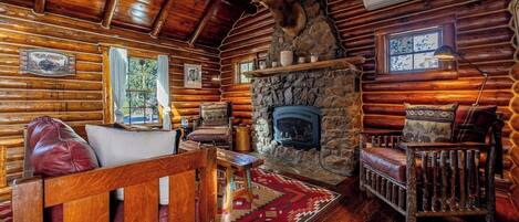 A log cabin interior with a stone fireplace, wooden furniture, and a mounted deer head.