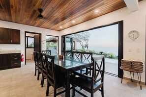 A modern kitchen with dark wooden cabinets, a glass dining table, and a view of the sea through large windows. - Casa Montaña - 5 Bedroom Ocean View Villa with Private Pool near Dominical. (Dominical)