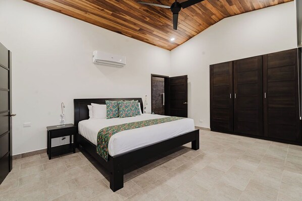A bedroom with a wooden ceiling, a bed with a white mattress and floral bedspread, a nightstand, a wardrobe, and a door. - Casa Montaña - 5 Bedroom Ocean View Villa with Private Pool near Dominical. (Dominical)