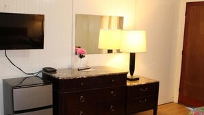 A hotel room with a dark wooden dresser, a small refrigerator, a flat-screen TV mounted on the wall, a bedside lamp, and a floral bedspread. - Royal Coach Motor Lodge (Hampton)
