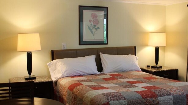 A hotel room with a bed, two bedside tables with lamps, a framed floral artwork on the wall, and a round table. - Royal Coach Motor Lodge (Hampton)