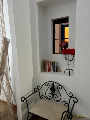 A white wall with a small window, a metal bench, and a bookshelf with books.