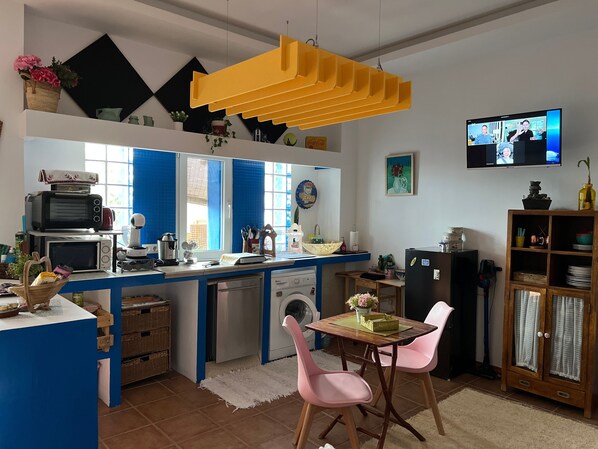 A kitchen with a blue countertop, a washing machine, a dining table with chairs, and a television displaying a video call.