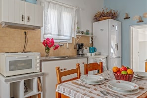 A compact kitchen with a microwave, kettle, and refrigerator. A dining table with plates and a fruit basket is set.