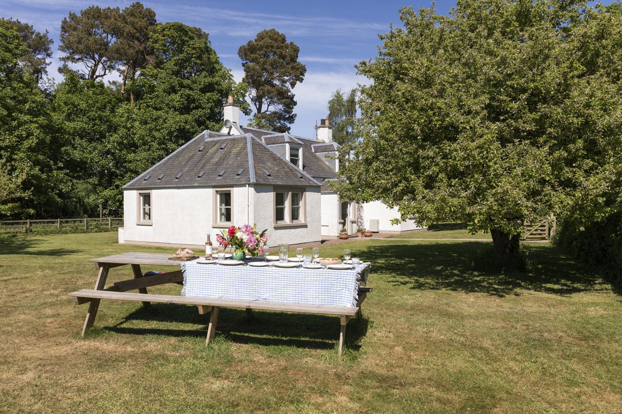 A white house with a pitched roof and a chimney, surrounded by trees and a picnic table set up on the lawn.