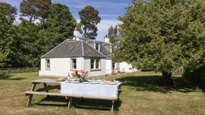 Una casa blanca con techo inclinado y chimenea, rodeada de árboles y una mesa de picnic en el césped.