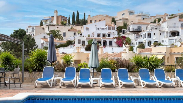 Ein Poolbereich mit Liegestühlen, einem Tisch und Blick auf weiße Gebäude mit Balkonen.
