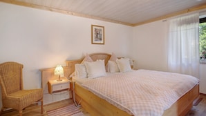 A bedroom with a wooden bed, a nightstand with a lamp, a wicker chair, and a framed picture on the wall. - Apartment 'Löwenzahn' with Mountain View, Terrace & Wi-Fi (Fischbachau)