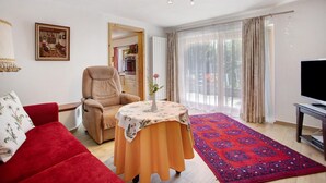 A living room with a red sofa, a beige armchair, a small table with a tablecloth, a television on a stand, and a large window with curtains. - Apartment 'Löwenzahn' with Mountain View, Terrace & Wi-Fi (Fischbachau)