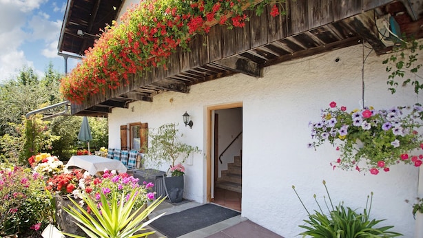 A building with a white exterior, a wooden balcony with hanging flowers, a door, and a staircase leading inside. - Apartment 'Löwenzahn' with Mountain View, Terrace & Wi-Fi (Fischbachau)
