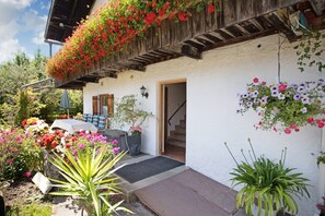 Een gebouw met een witte gevel, een houten balkon met bloemen en een trap die naar de ingang leidt.
