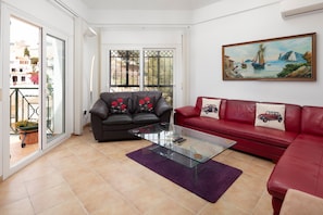 A living room with a glass coffee table, a black sofa with red floral cushions, a red sofa with a car pillow, and a painting of a coastal scene. - Villa Miramar Burriana Beach 6 pax with Sea Views & Pool (Nerja)