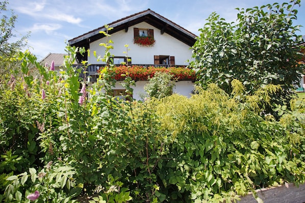 A traditional chalet with a balcony, surrounded by lush greenery and flowering plants.
