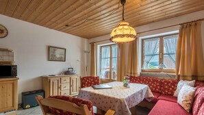 Een gezellige kamer met een houten plafond, een eettafel met een tafelkleed met dessin, een bank met decoratieve kussens en een raam met gordijnen.