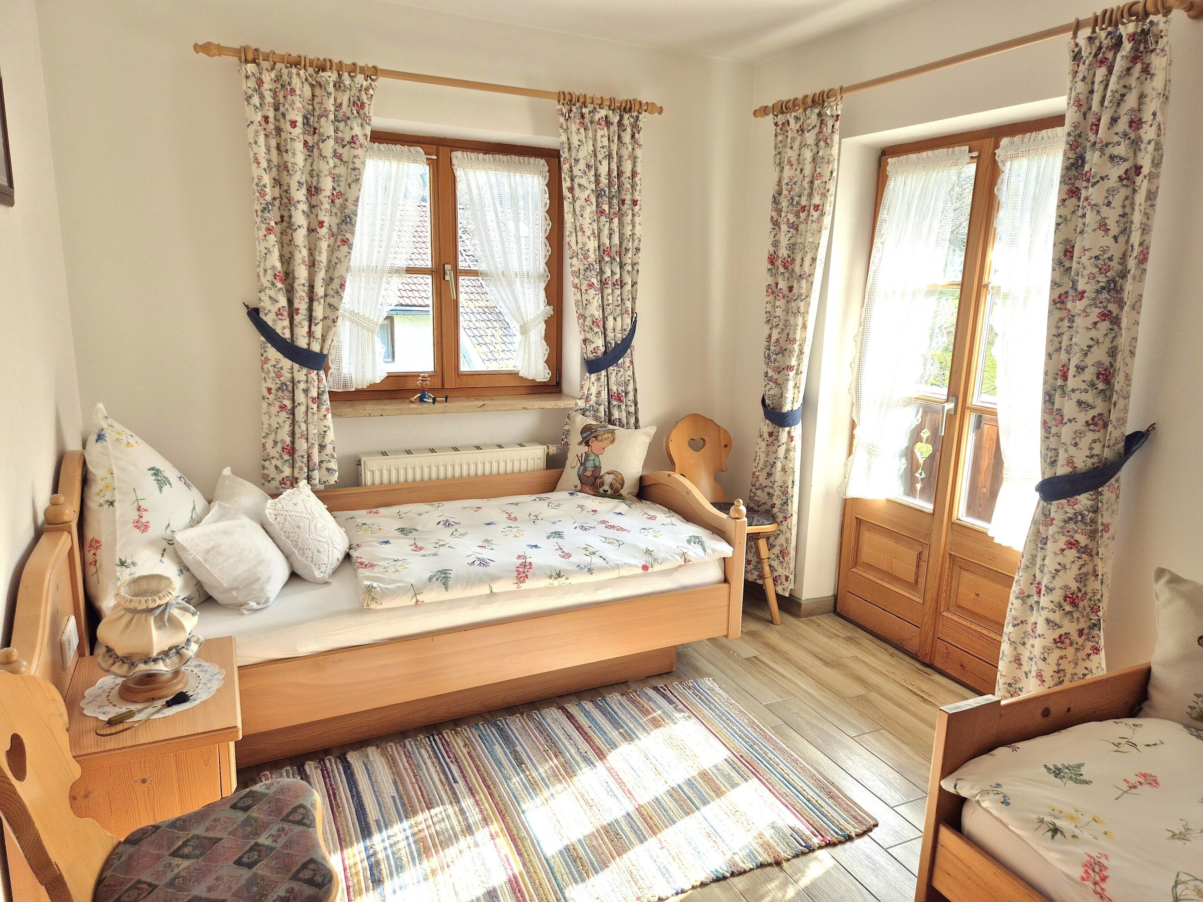 A bedroom with a wooden bed, floral curtains, a wooden chair, a bedside table with a lamp, and a window with a view.