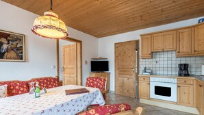 Een kamer met een eettafel, stoelen, een televisie, houten kasten en een betegeld keukenhoekje.