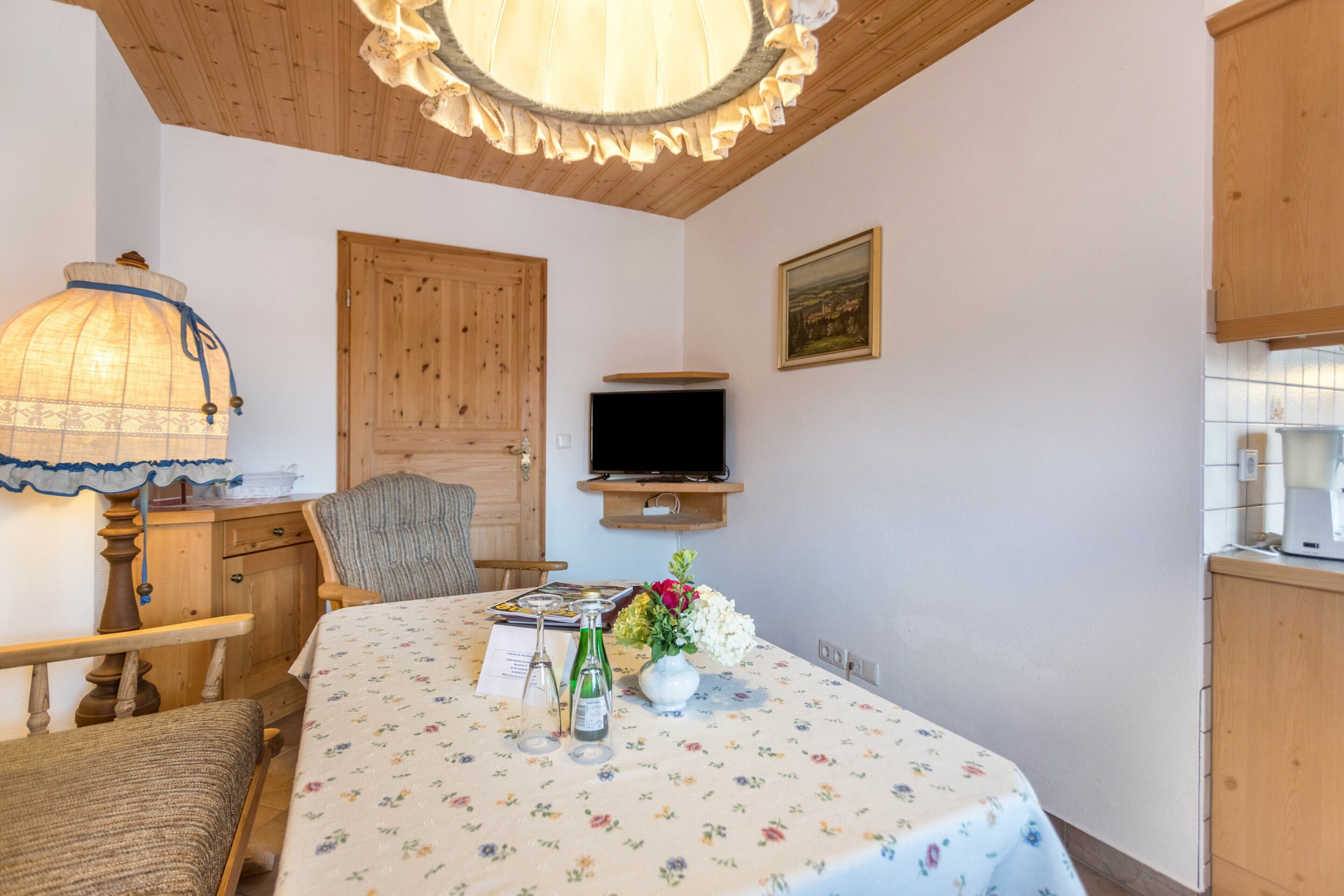 Ein Zimmer mit Holzboden, einem Tisch mit einer blumigen Tischdecke, einem Stuhl, einer Lampe, einem Fernseher und einem Schrank.