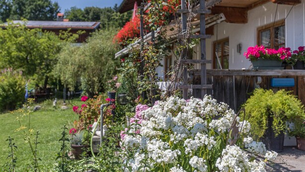 A traditional wooden house with a balcony, surrounded by lush greenery and flowering plants. - Apartment 'Enzian' with Mountain View, Balcony and Wi-Fi (Fischbachau)