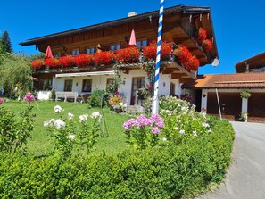 A traditional wooden building with a balcony adorned with red flowers, a well-maintained garden with various plants, and a clear blue sky.
