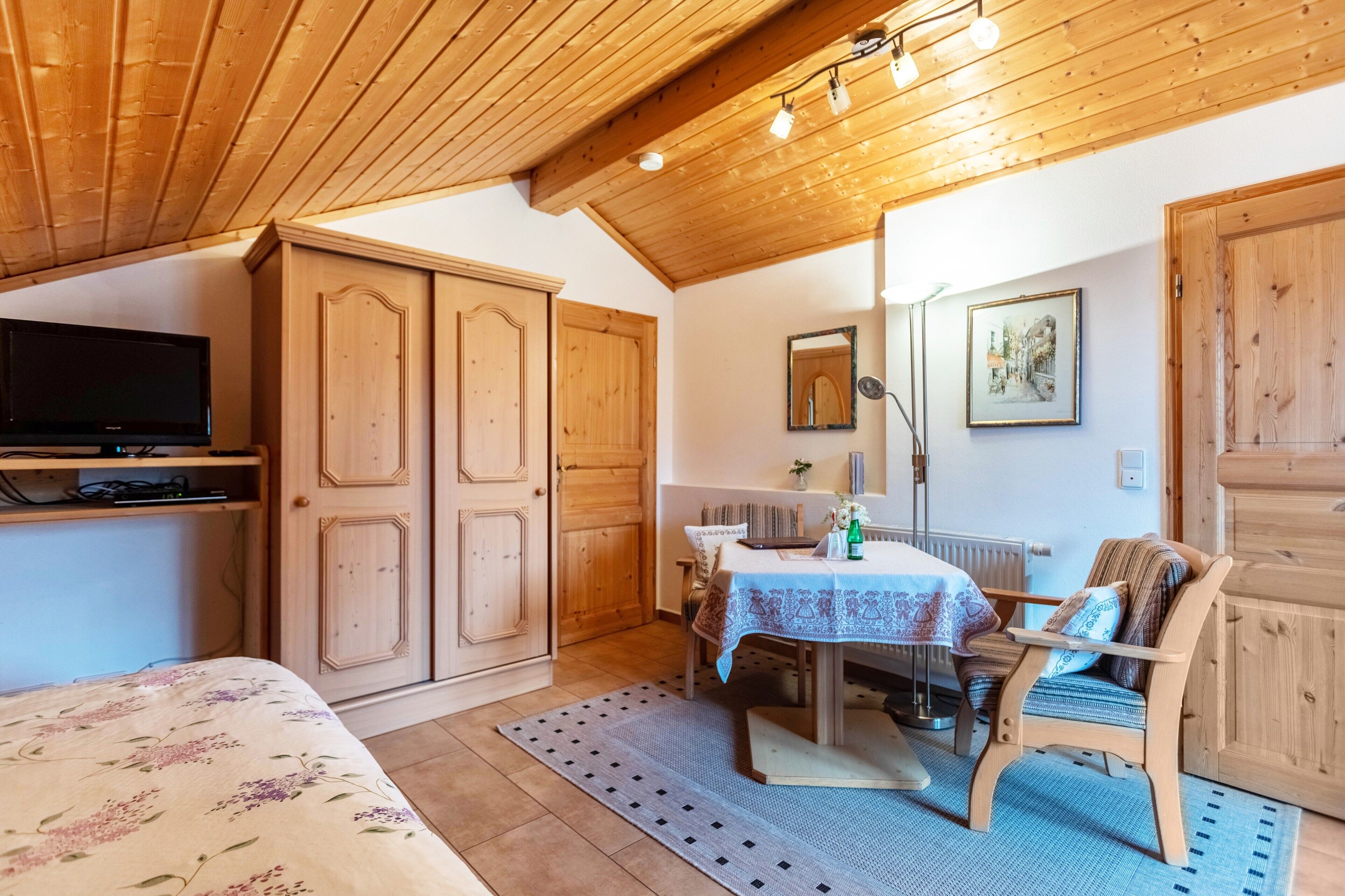 Une pièce comprenant un lit, une table recouverte d’une nappe, une chaise, des meubles en bois, un téléviseur et un miroir au mur.