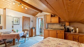 Una habitación acogedora con cama, una mesa, una silla, un pequeño rincón de cocina y un ventanal con cortinas.