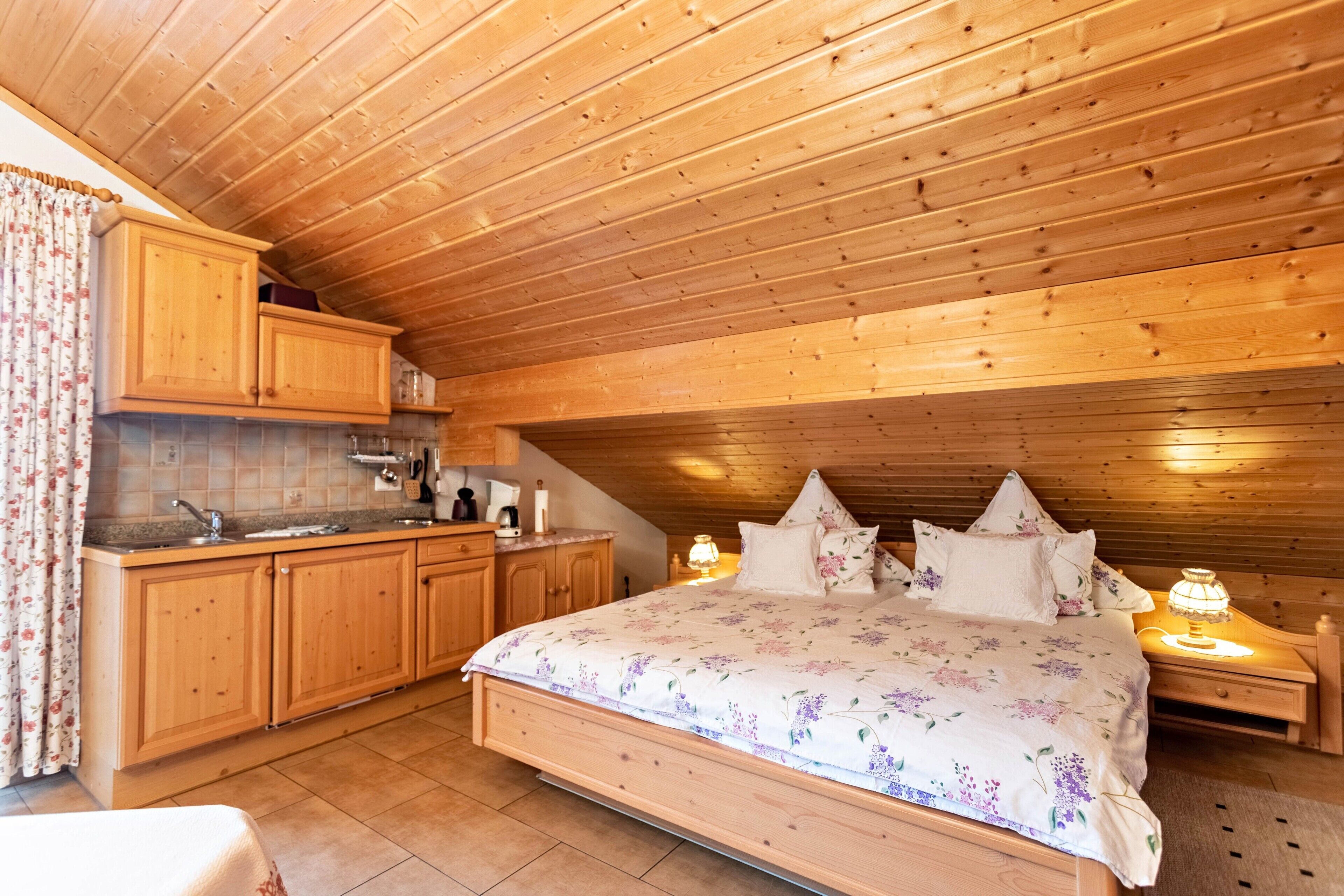Une chambre avec un plafond en bois, un grand lit avec une literie à motifs floraux, une table de chevet avec une lampe, et un coin cuisine avec des placards en bois et un évier.