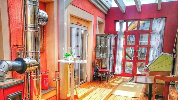 Una habitación con una pared roja, un vidrio, una mesa pequeña con una planta y una silla de madera.