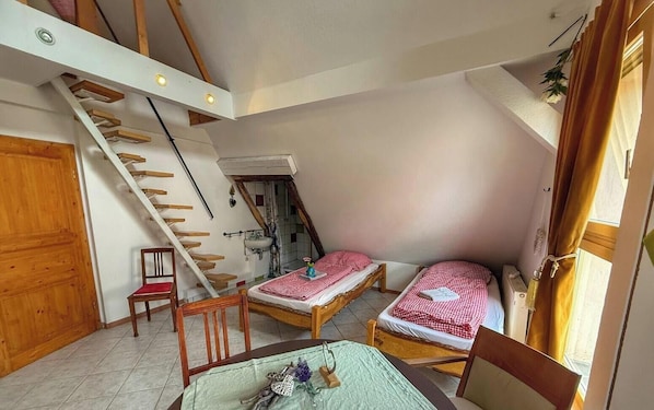A compact room with two beds, a wooden chair, a dining table, a staircase, and a small bathroom.