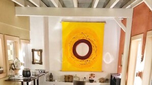 Una larga mesa de comedor dispuesta para una comida, sillas, un cuadro abstracto amarillo en la pared y una cocina visible al fondo.