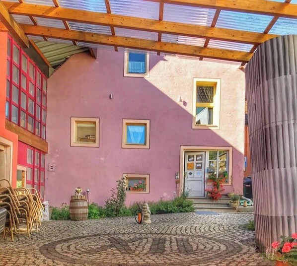 Un bâtiment rose doté d’un patio couvert, d’une porte en bois et d’un petit jardin.