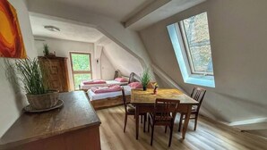 A room with a wooden table and chairs, a bed with pink bedding, a window, and a potted plant.