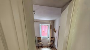 Ein schmaler Flur mit Holzboden, der zu einem Zimmer mit Fenster, zwei Stühlen und einem Heizkörper führt.