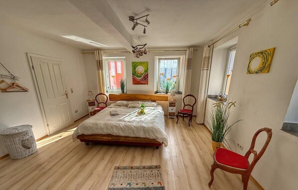 Une chambre à coucher avec un grand lit, des tables de chevet, une chaise, un luminaire suspendu et une fenêtre avec des rideaux.