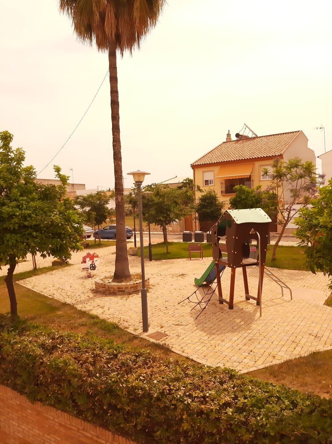 Un parque infantil con tobogán y columpios, rodeado de árboles y un edificio al fondo.