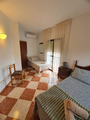 A room with a checkered floor, a bed with a patterned bedspread, a chair, a wooden nightstand, and a window with curtains.