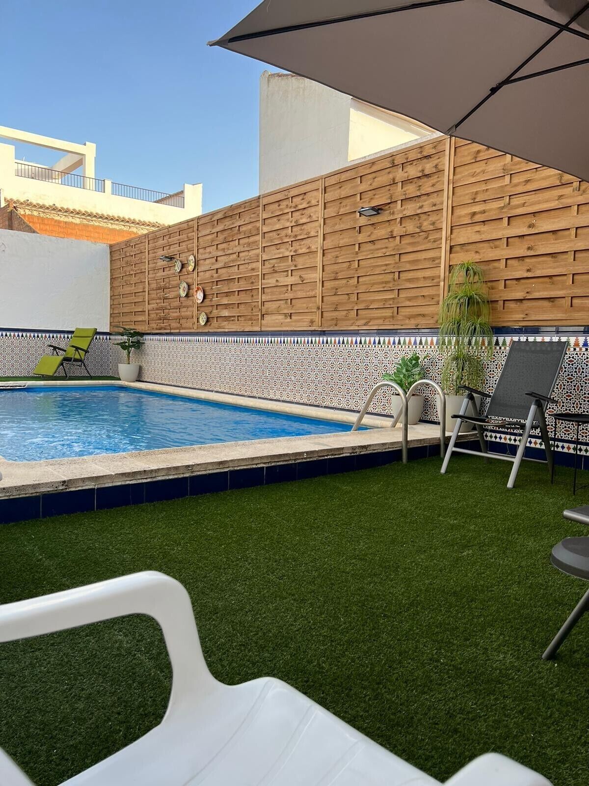 Una piscina con cerramiento de madera, un patio con sillas y un pequeño jardín.