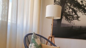 Um quarto com mesa de vidro, cadeira de vime com almofadas, luminária de chão e um quadro na parede.