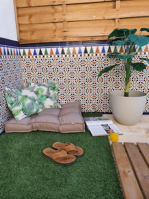 A small outdoor seating area with a sofa, a large potted plant, and a pair of sandals on artificial grass.
