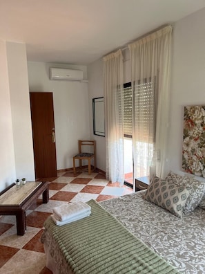 A bedroom with a checkered floor, a bed with patterned bedding, a chair, a small table, and a painting on the wall.