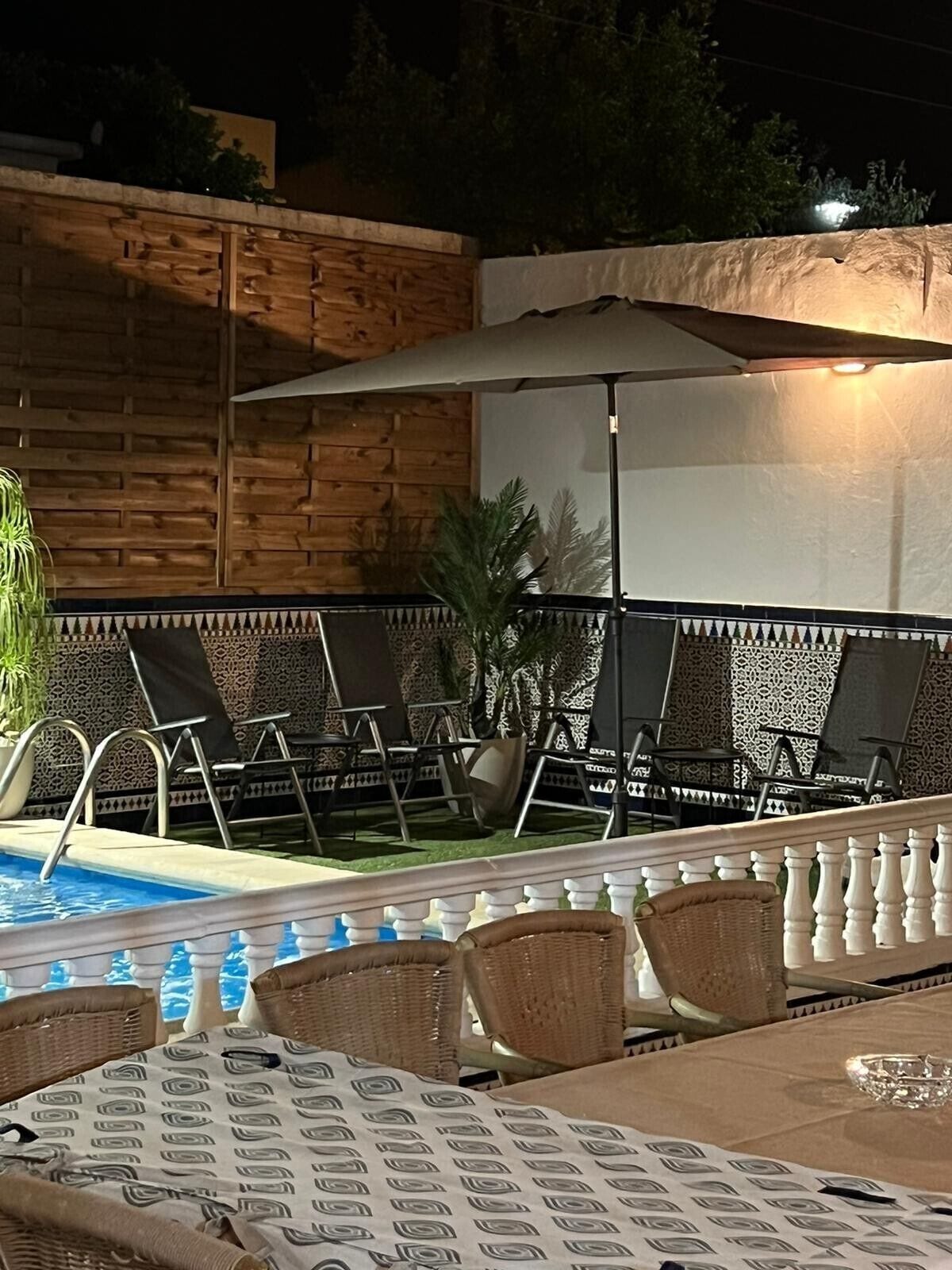 An outdoor pool area with lounge chairs and a table, surrounded by a wooden wall and patterned tiles.