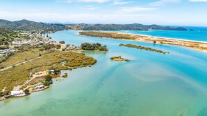 An aerial view of a coastal area with clear waters, a beach, and a small settlement with buildings and greenery. - Stylish Waterfront Studio Sanctuary! (Ngunguru)