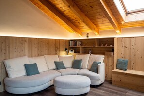 Une pièce chaleureuse avec un canapé d’angle, un petit pouf, des murs en bois, un plafond en pente avec des poutres apparentes et une étagère remplie de livres et d’objets décoratifs.