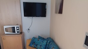 Una stanza piccola con un divano letto, una televisione a muro, un forno a microonde su un mobile e una sedia.